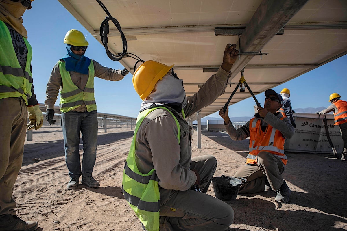 Workers in safety gear install solar panels in a sandy desert environment