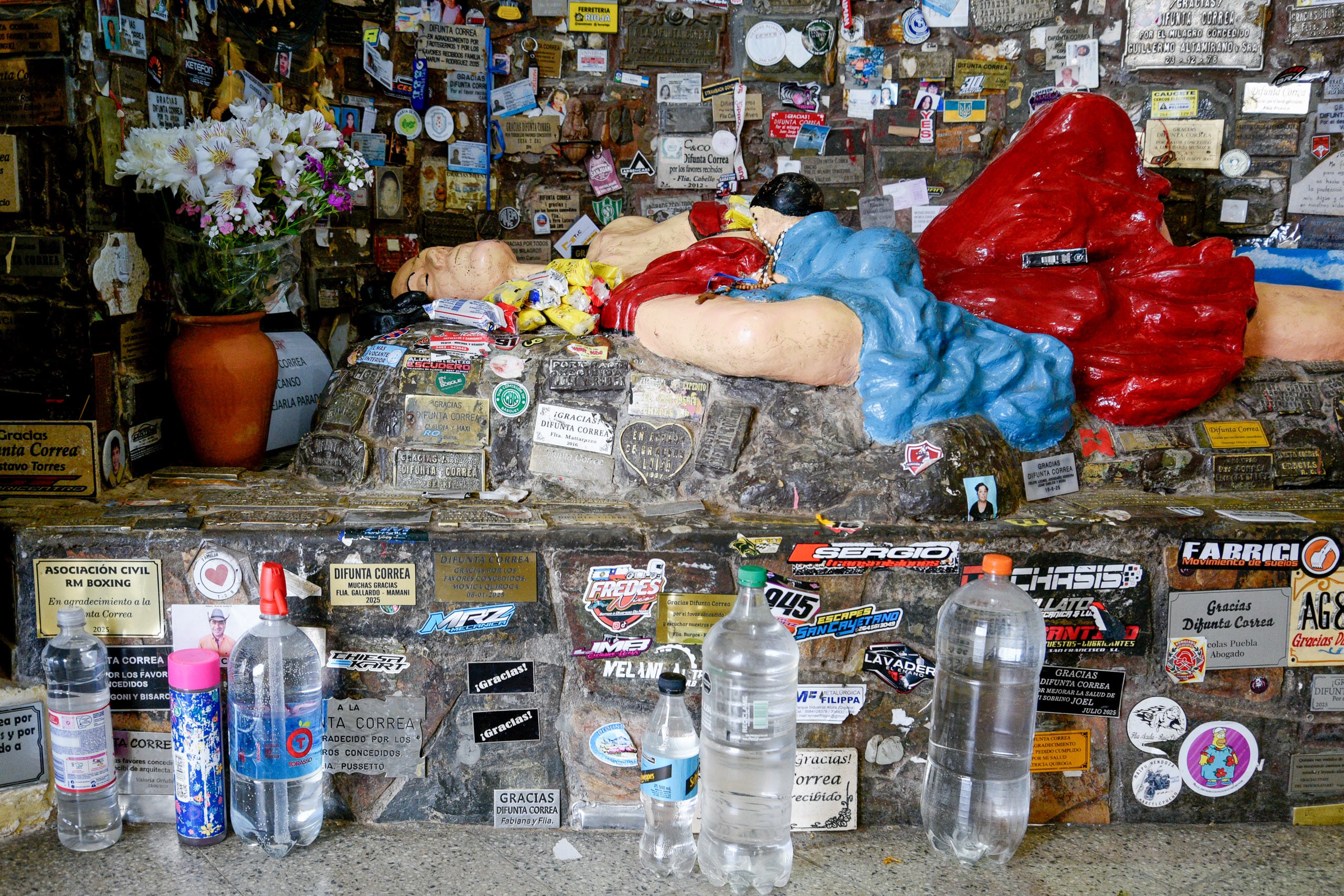 Una estatua de una mujer rodeada de diversas pegatinas y ofrendas, con flores y botellas de agua colocadas delante