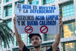 A person holds a sign at a protest stating "The water is ours and we defend it! We want clean, good-quality water!"