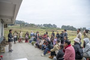 Villagers listening to a speaker outdoors, with fields and hills in the background