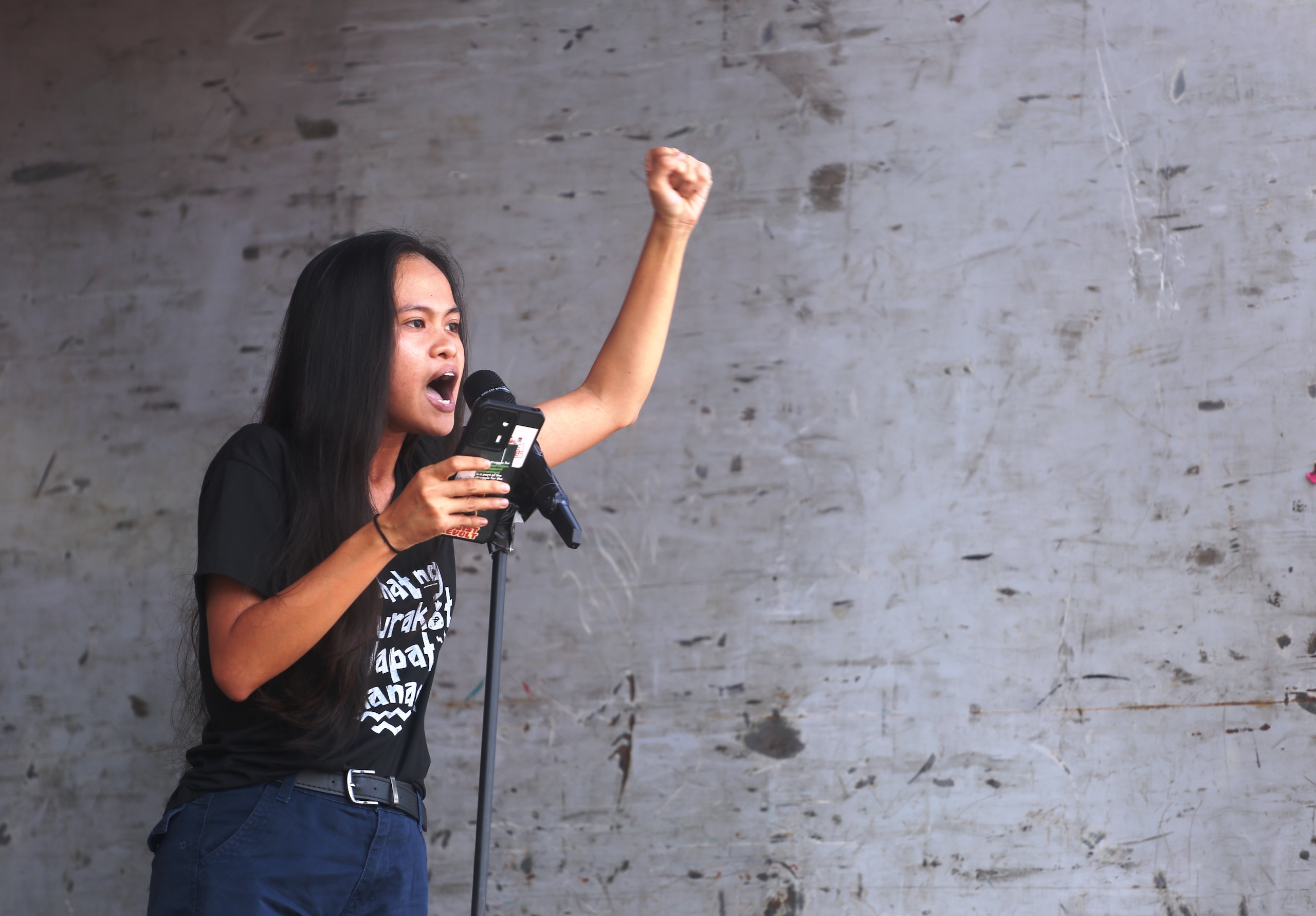 Jonila Castro stands at a microphone, raising her fist while speaking