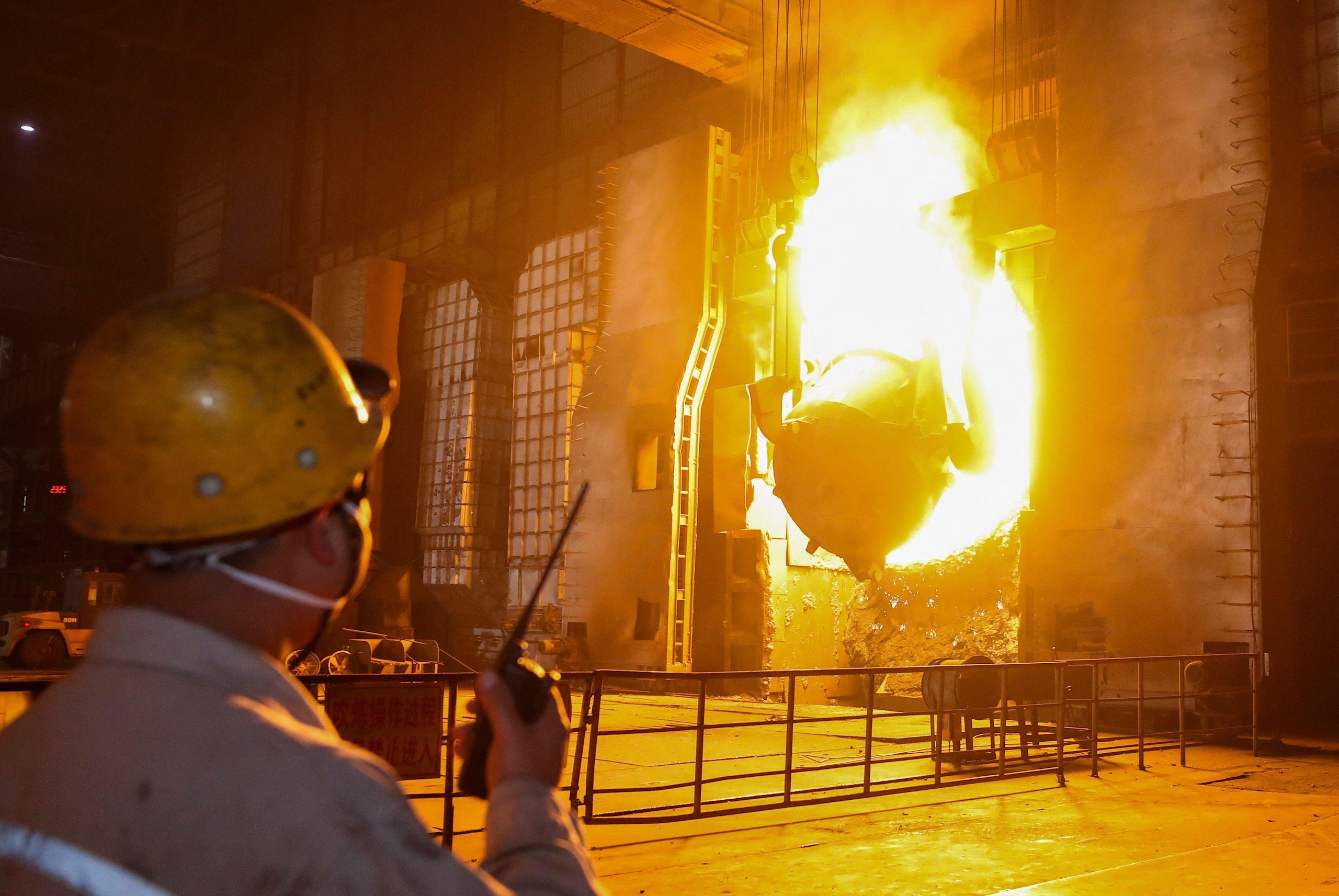 A worker in a hard hat holds a walkie-talkie inside a steel mill. A large furnace glows brightly, casting an intense orange light.