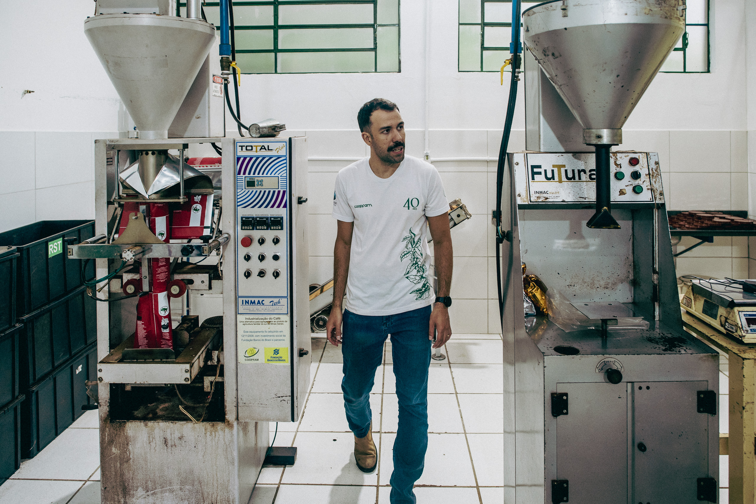 Man in a white t-shirt walks between industrial coffee packaging machines in a factory setting