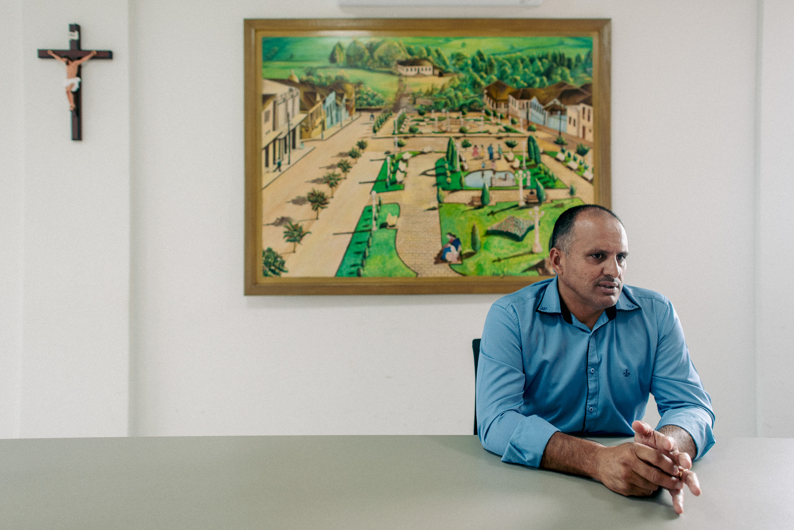 Man in blue shirt sitting at a table with a landscape painting on the wall behind him.