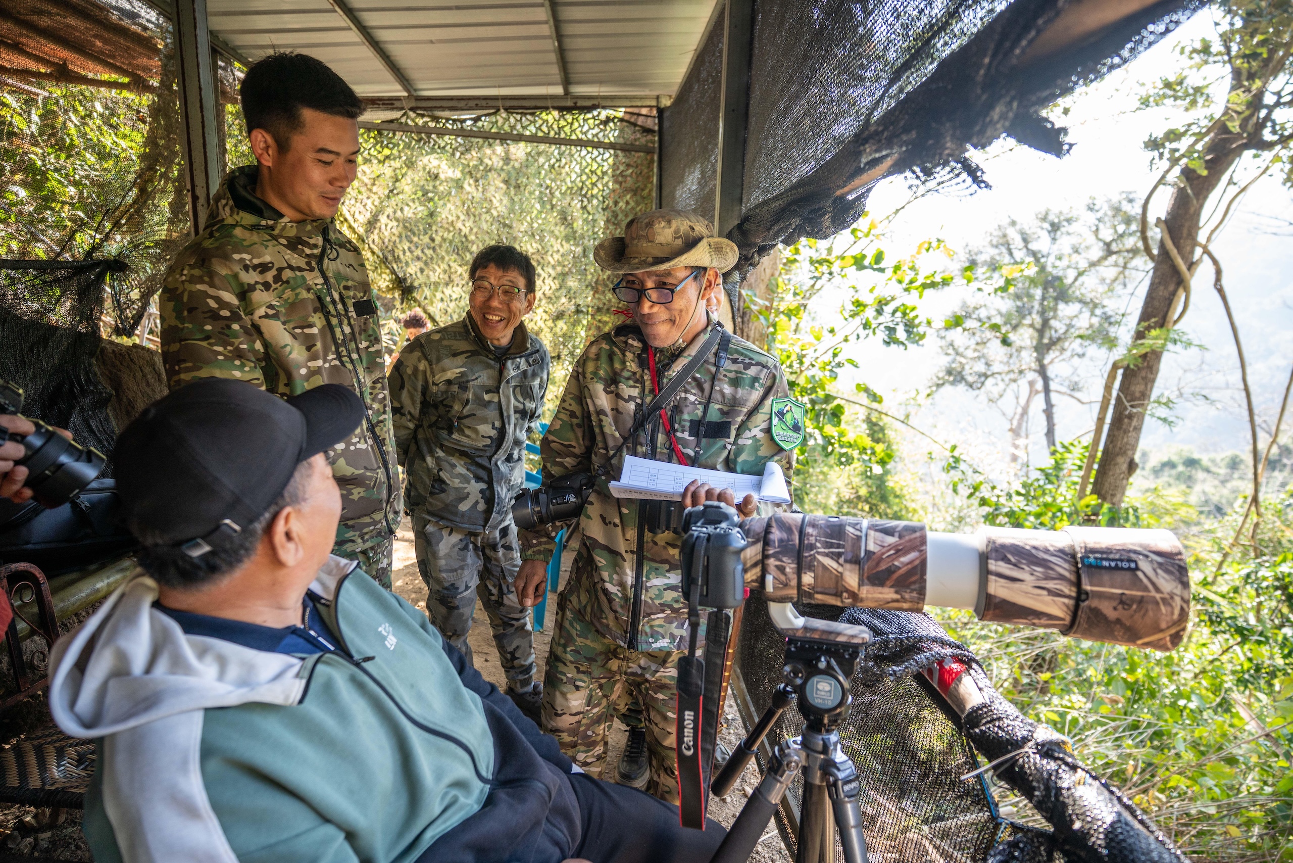 Four people in camouflage attire are in a wooded observation shelter