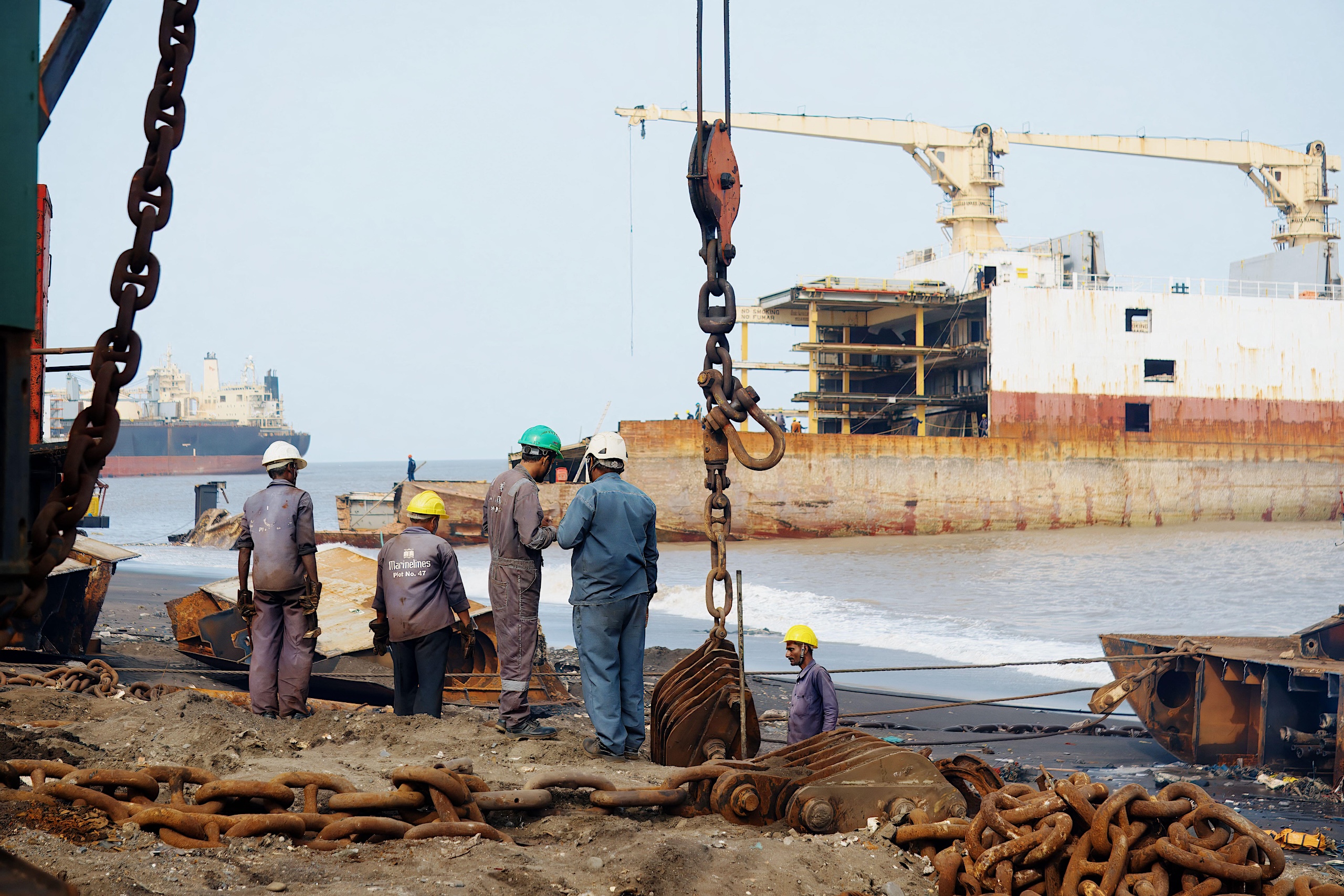 Workers in helmets and uniforms at a shipbreaking site, with a rusted ship in the background