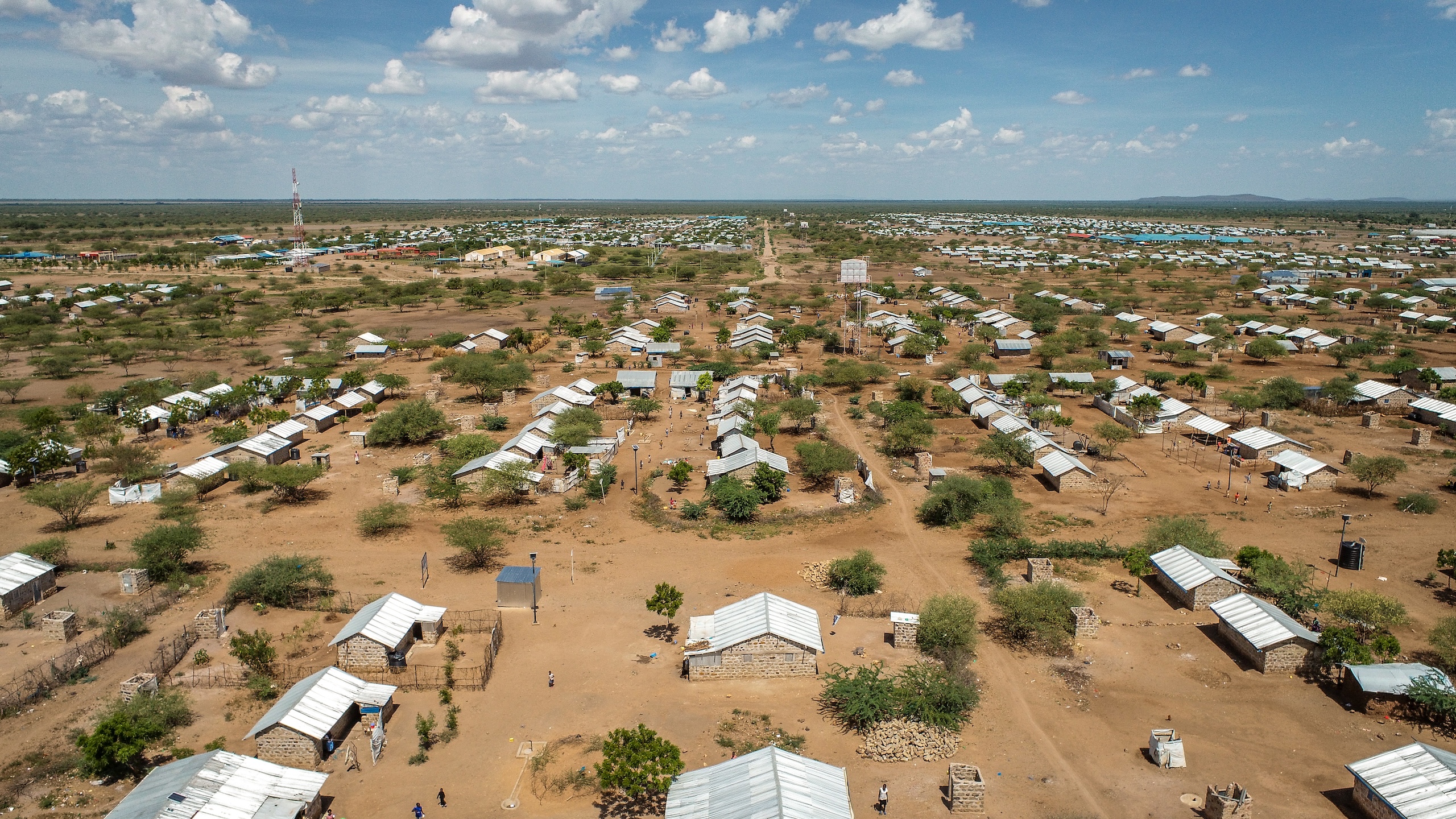 Aerial view of a vast refugee camp, with numerous tin-roofed shelters scattered across a dry, brown landscape dotted with sparse trees