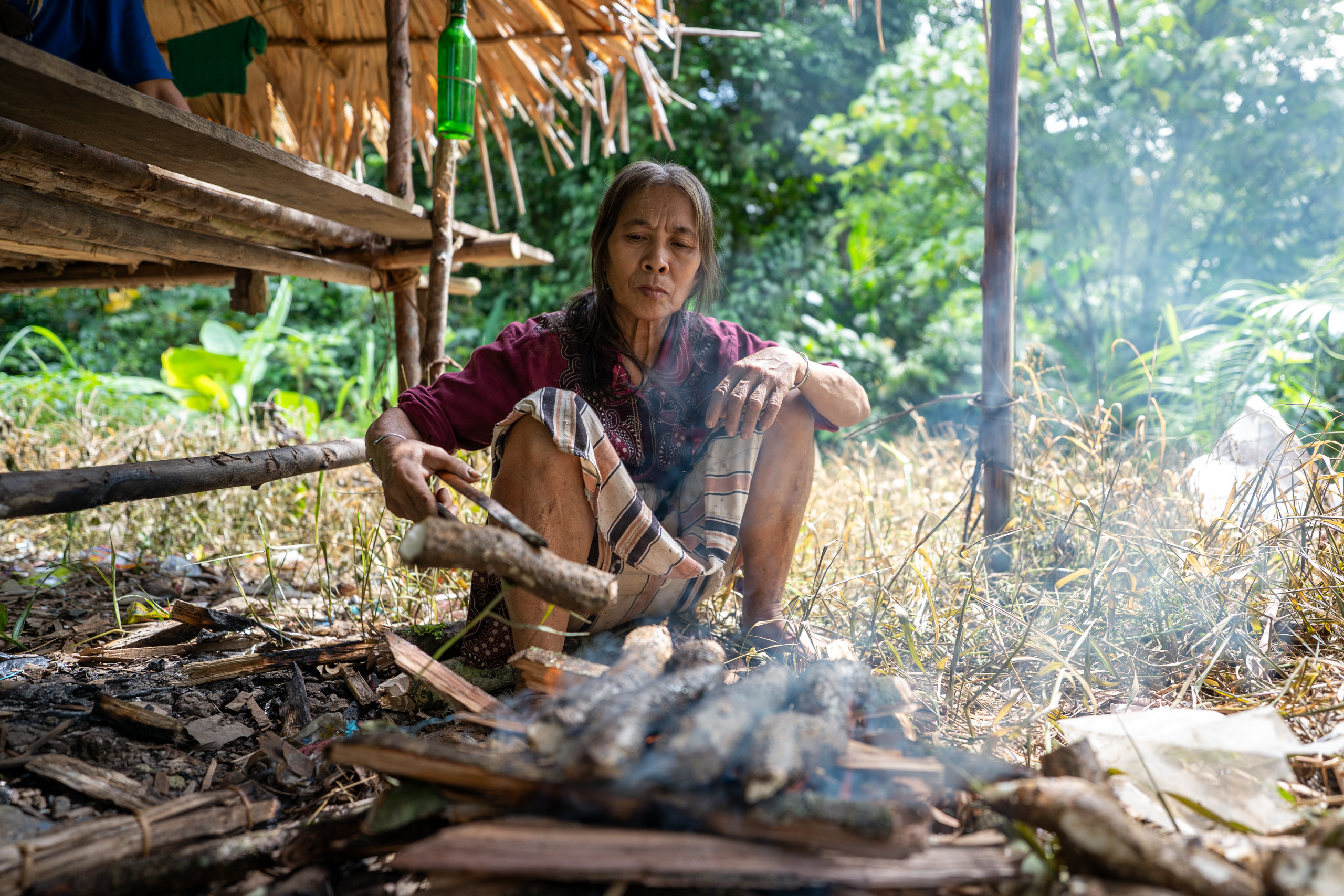 A woman roasts plant tubers over an open fire in front of a small hut