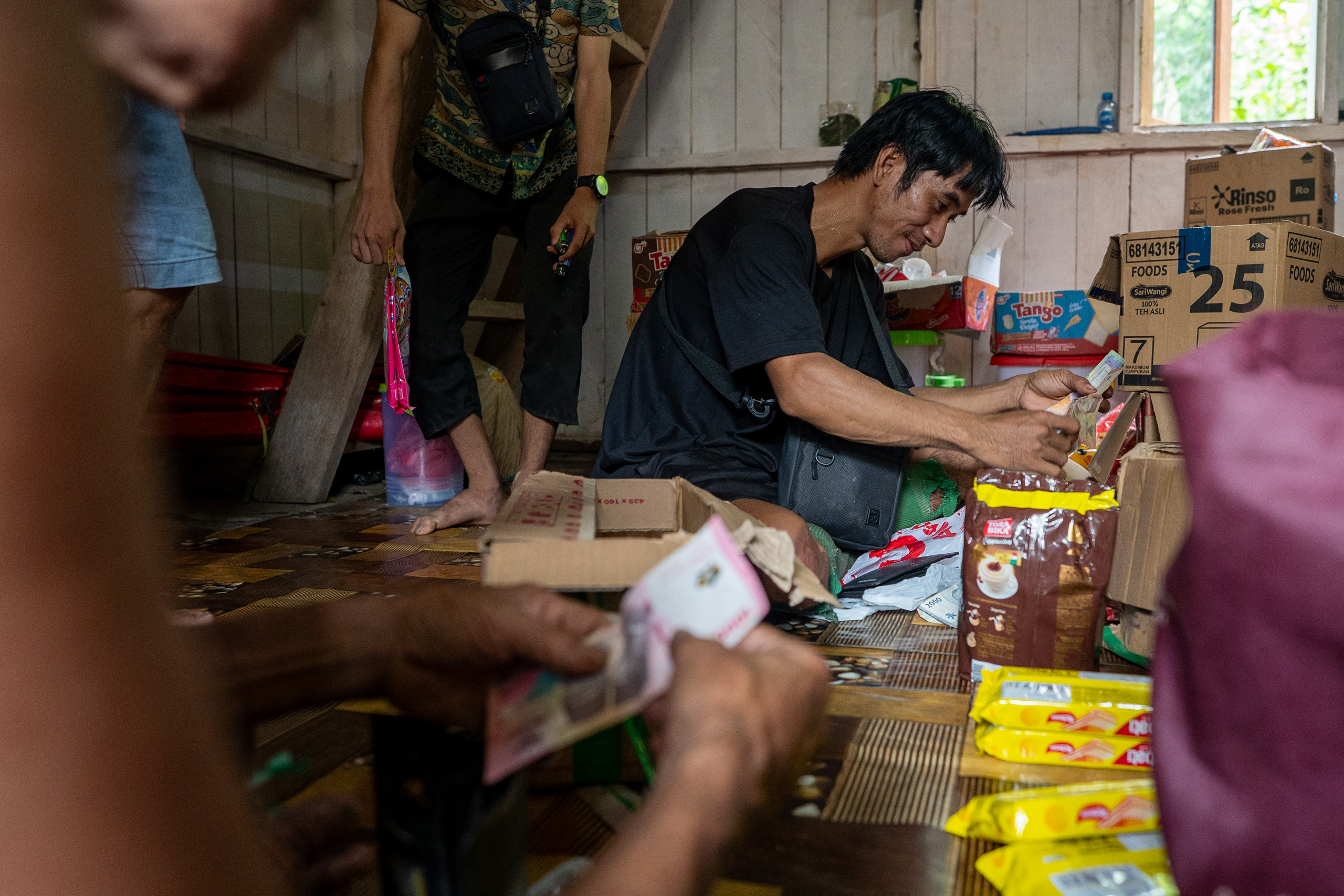 A man sits on a floor sorting money, surrounded by various food packages and cardboard boxes
