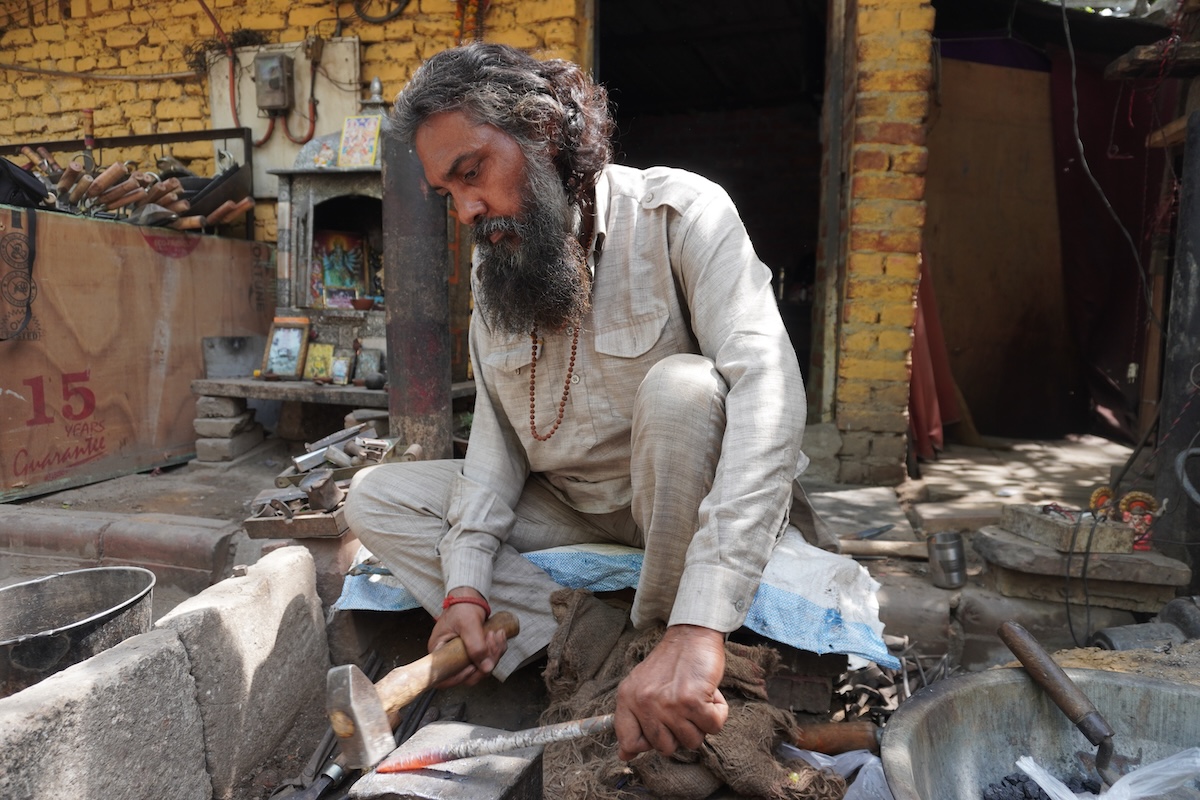 A man sits on the ground and working with a hammer and chisel on a metal rod