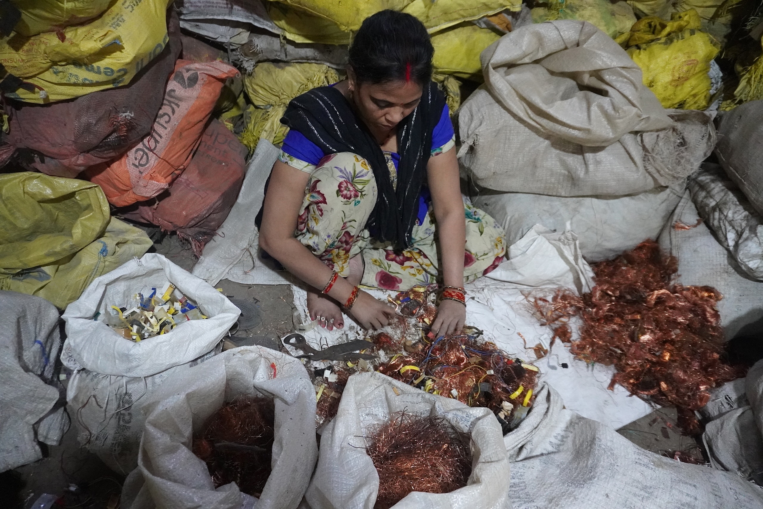 A woman sits sorting copper wires amidst large bags of recyclables