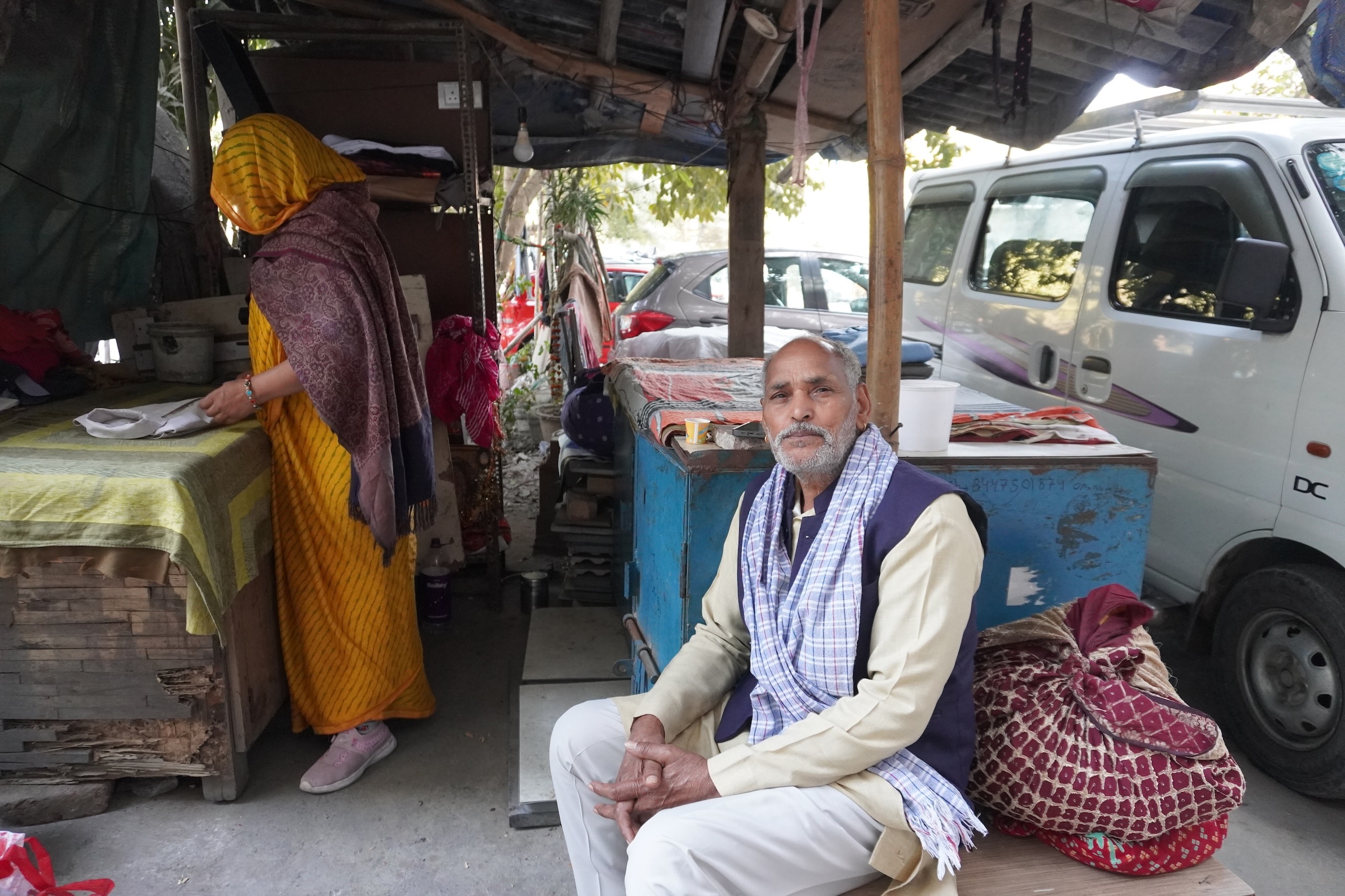 A man sits on a bench outside a workshop, with a woman irons clothes in the background 