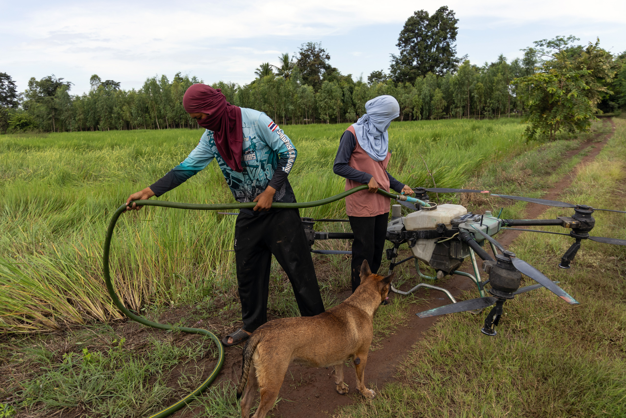 Two people wearing head coverings fill an agricultural drone with liquid from a green hose in a field