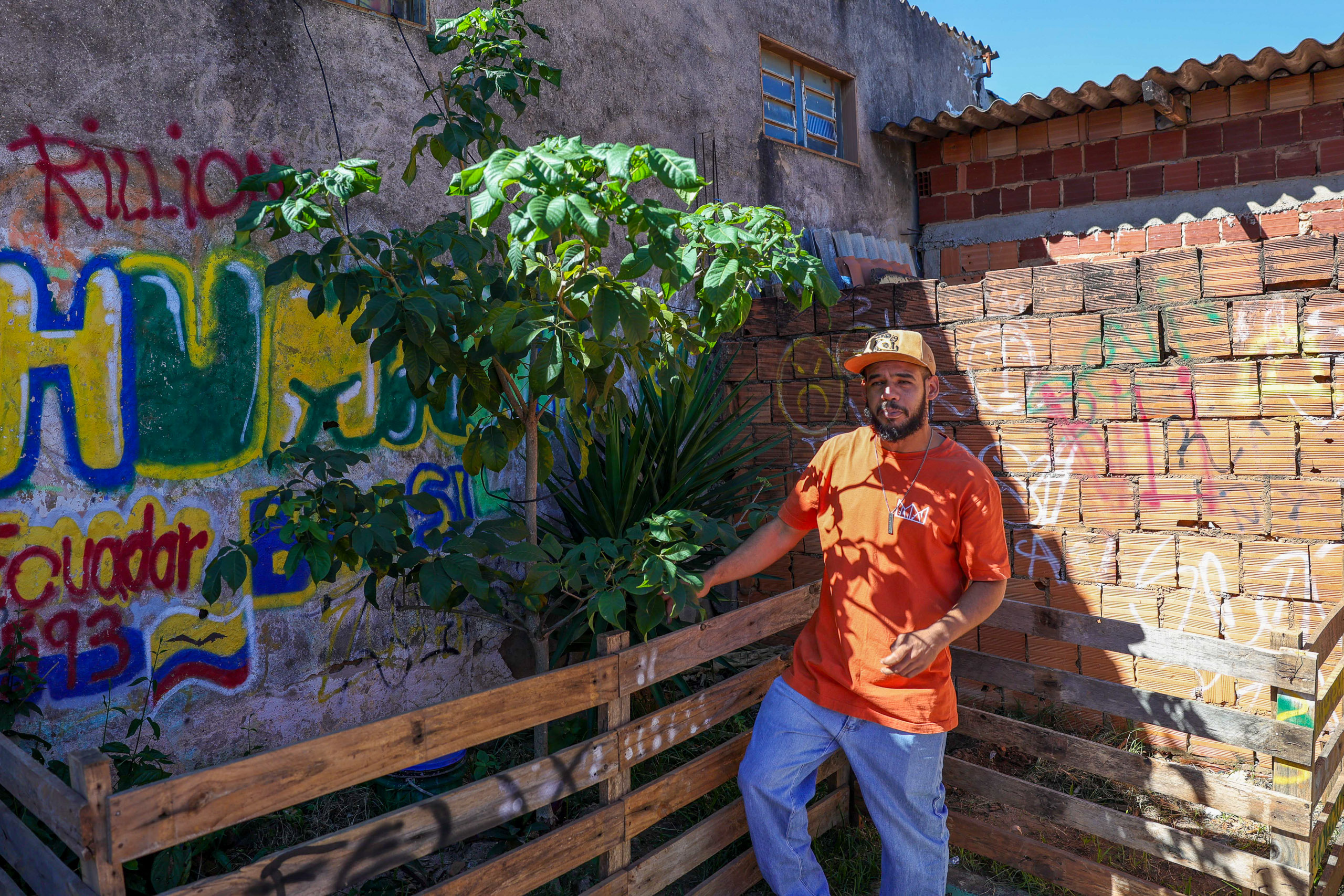 Man in orange shirt standing next to a tree with a graffiti wall in the background