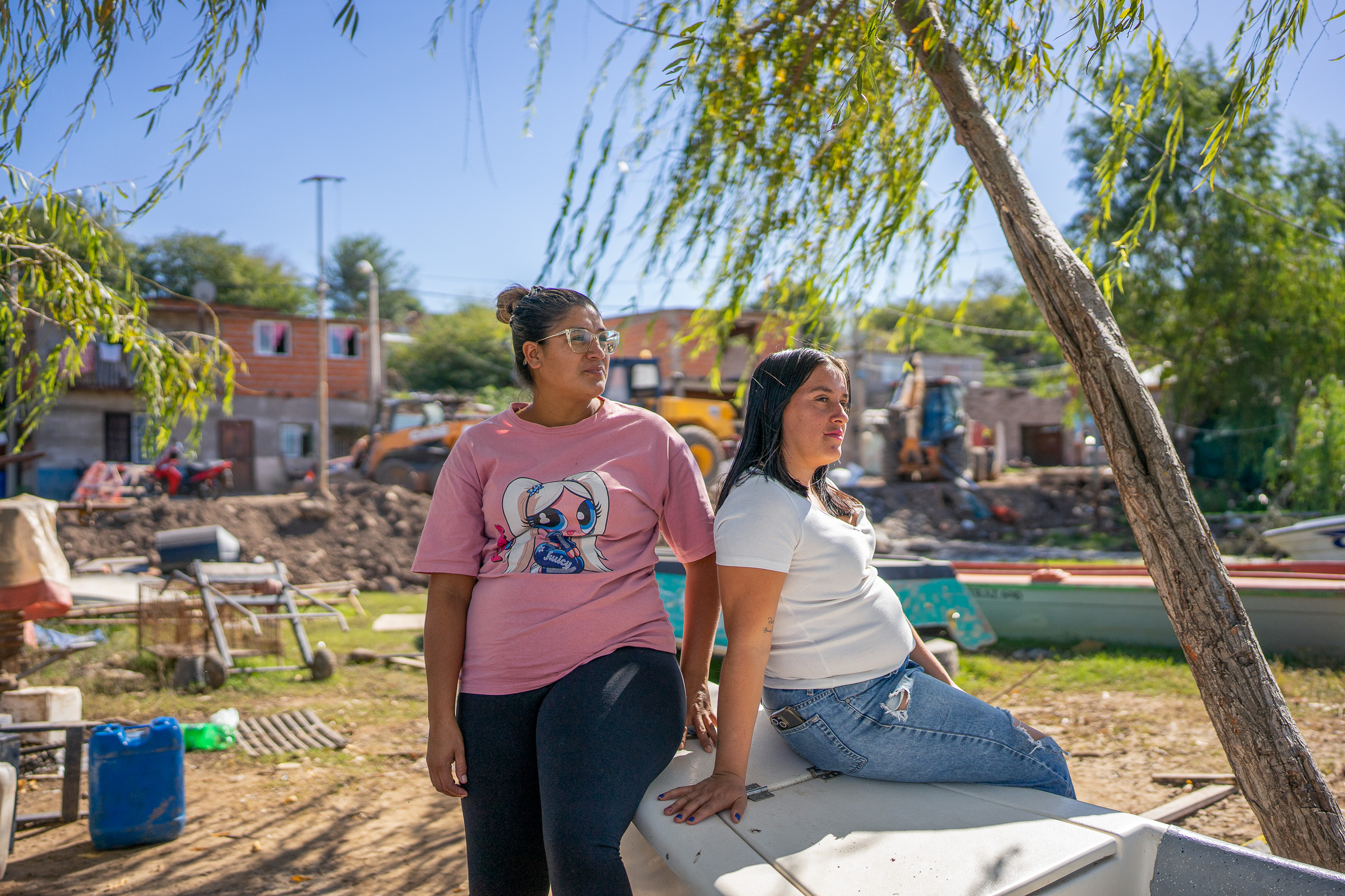 Dos mujeres de pie bajo un árbol, con casas al fondo