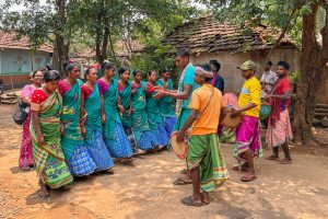 A group of women in green and blue traditional saris dance in a line in a rural village setting.