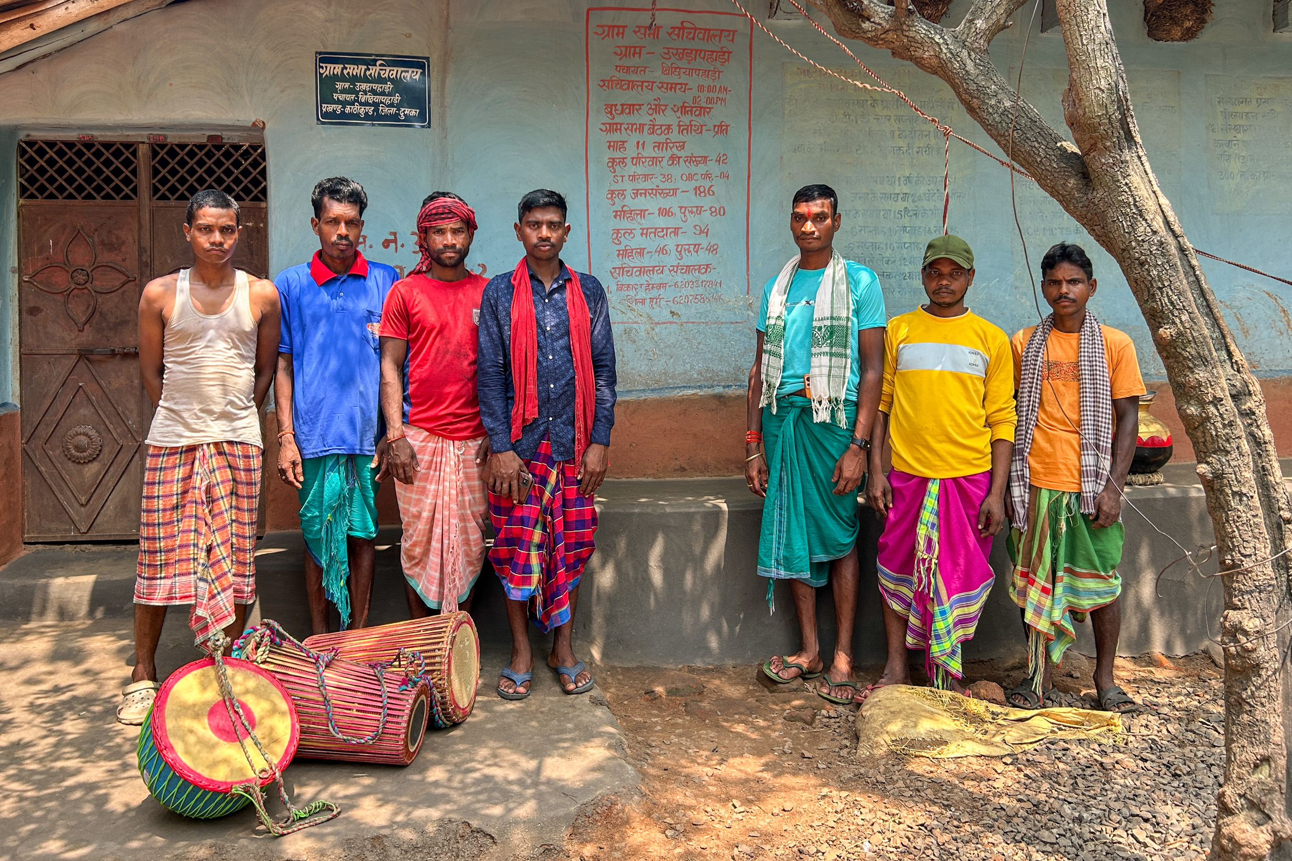 Seven men wearing traditional lungis and shirts stand in front of a building , with three large drums placed on the ground nearby.
