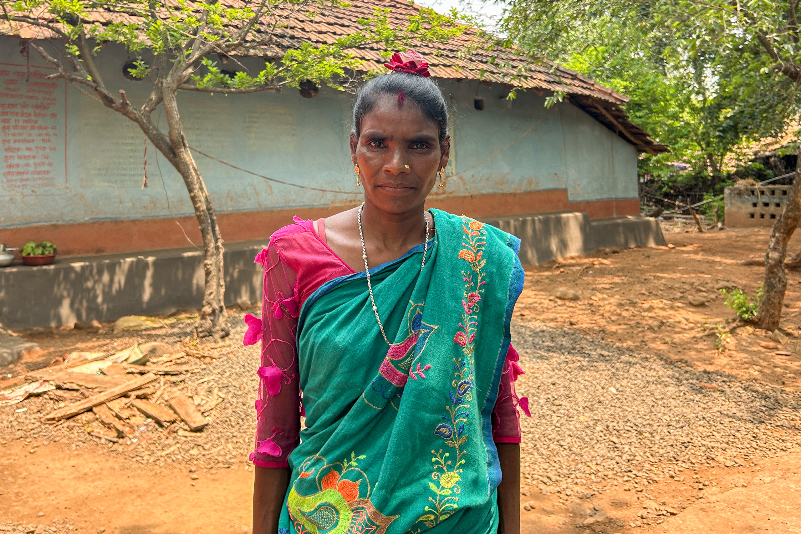A woman in green and blue traditional saris  standing in front of a rural village home.