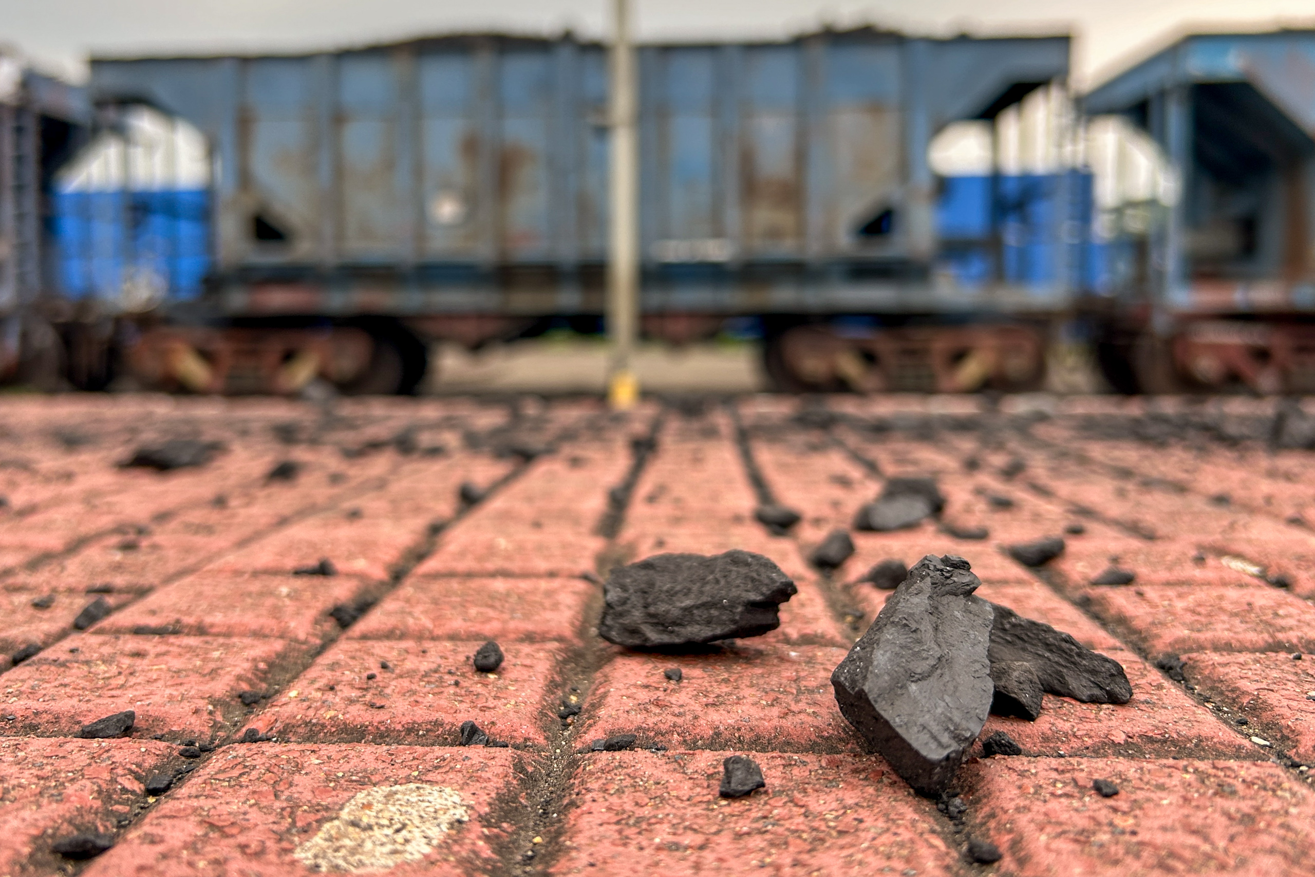 Close-up of black coal chunks scattered on a red brick ground, with industrial freight train cars in the background.