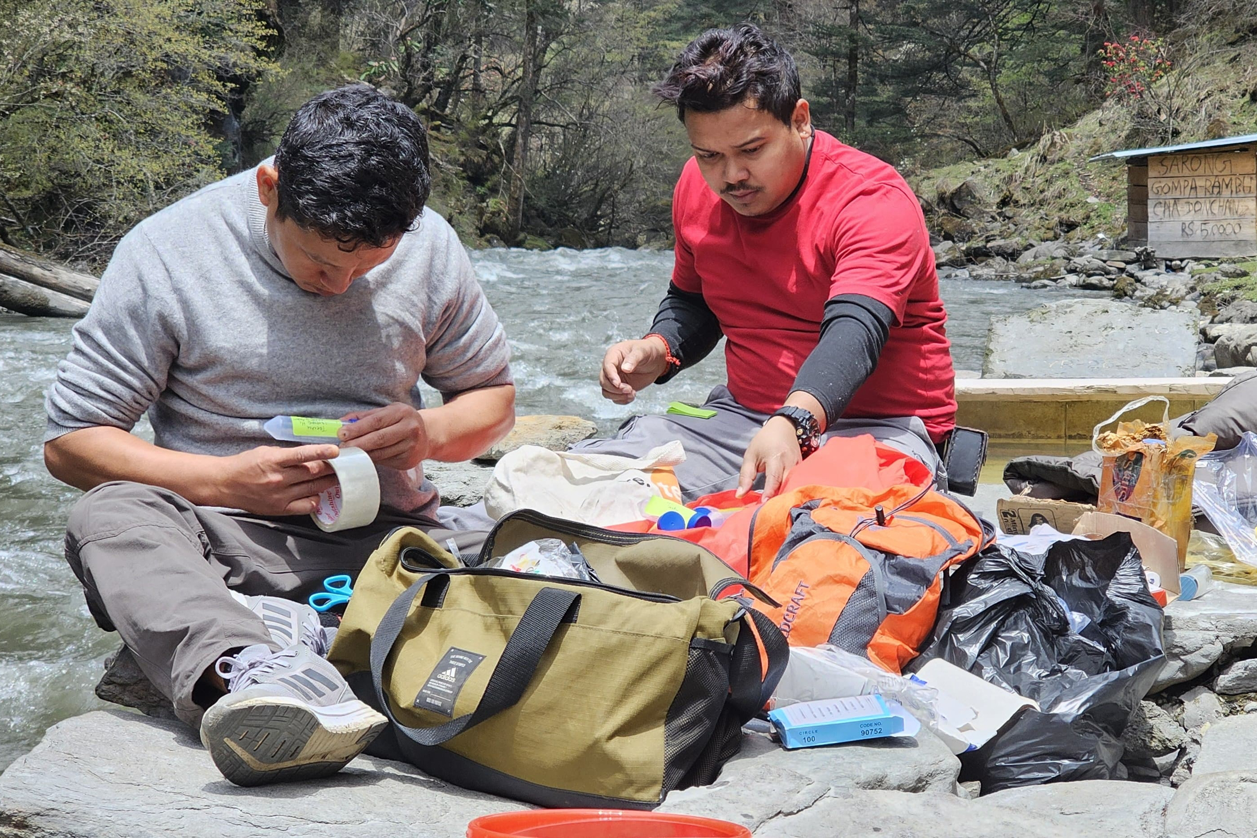 two men sitting on rocks near water and surrounded by equipment 