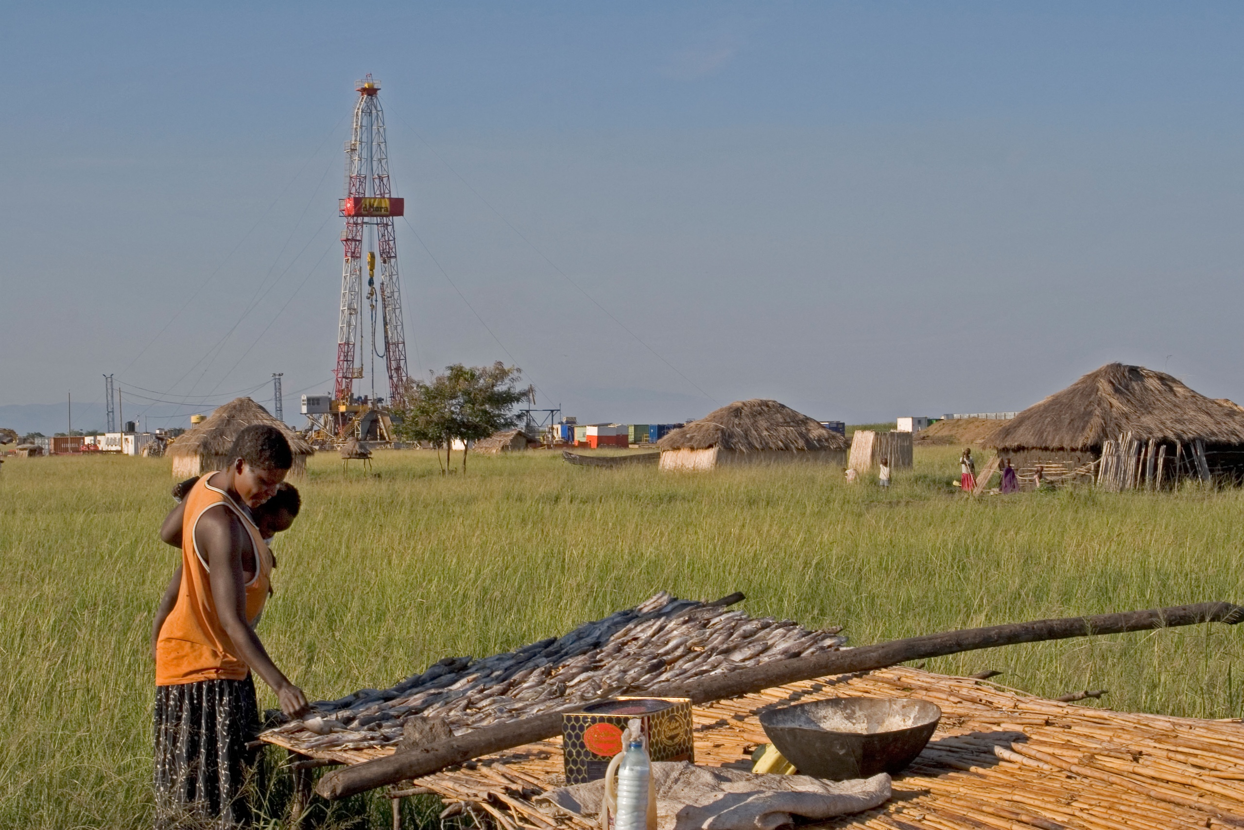 Woman drying fish on mat in rural village with an oil exploration rig in the background
