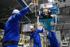 Three workers in blue uniforms are installing batteries in an EV production line
