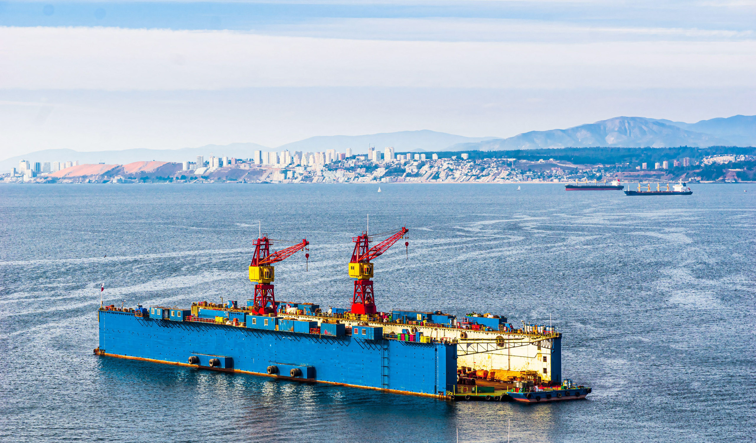A blue barge with two red cranes and yellow tops is floating on calm water, with a coastal city and hills in the background.

