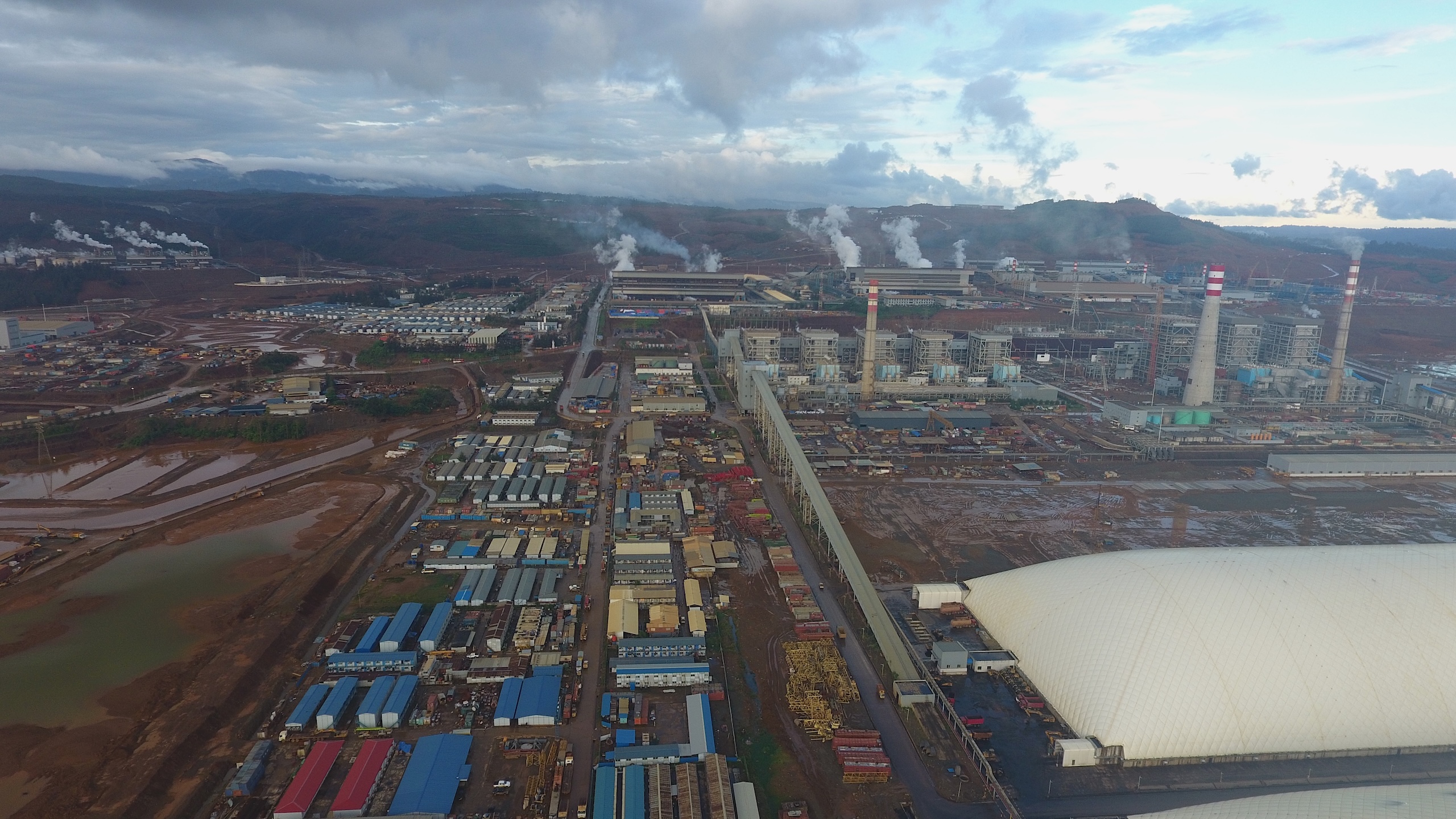 Aerial view of a large industrial complex with factory buildings, tall smokestacks emitting steam, and rows of small structures under a cloudy sky.