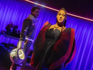 A stage set of a woman with red pleated sleeves stands before a man in a space suit holding a helmet
