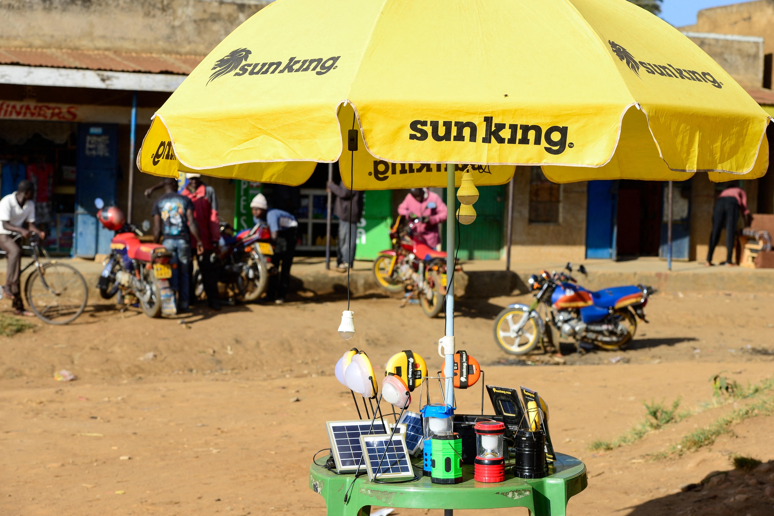 A yellow Sun King umbrella shades a green table displaying various solar-powered lamps and small solar panels in an outdoor market setting.