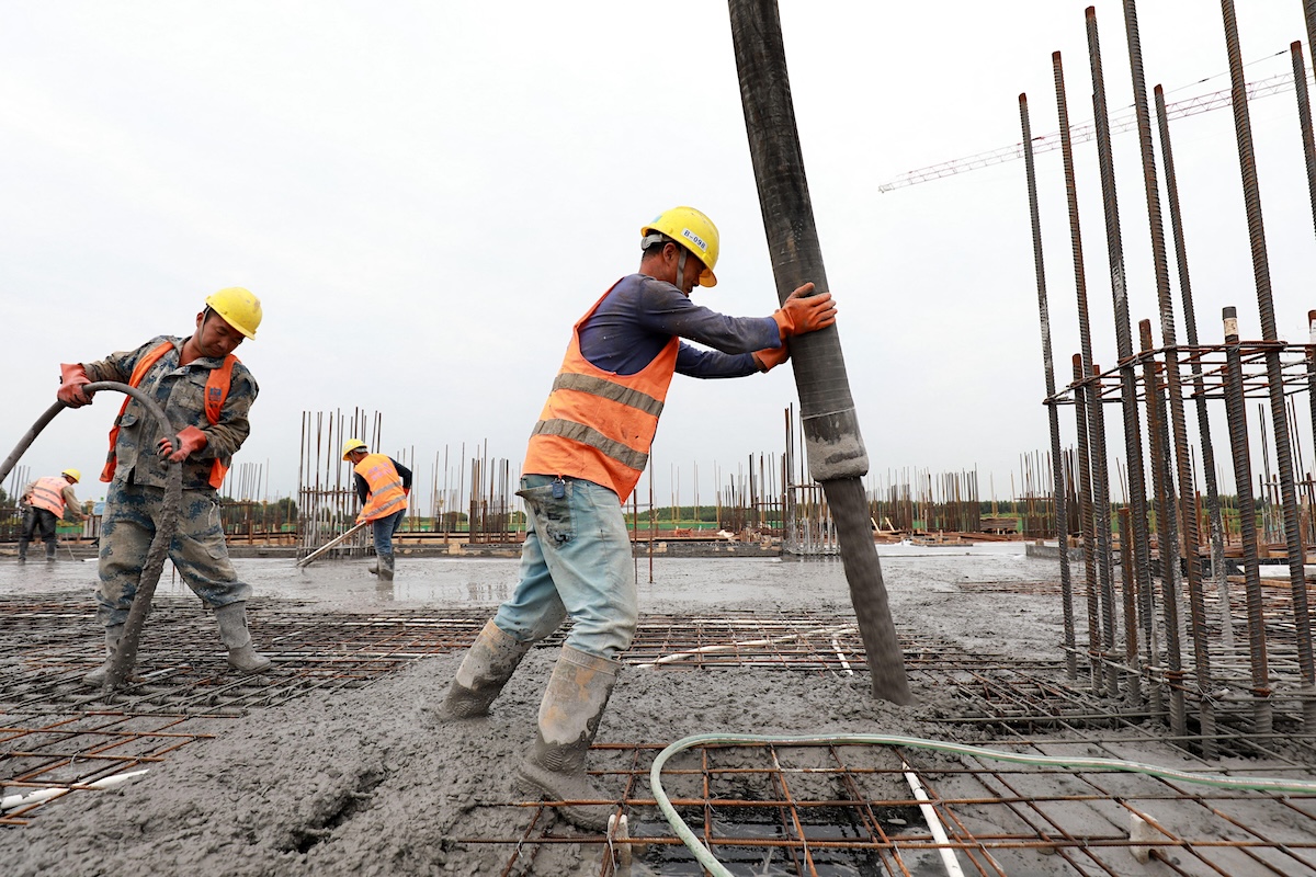 Construction workers pour concrete from a large hose onto a steel rebar grid at a building site