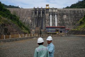 Two workers in hard hats stand in front of a large concrete dam