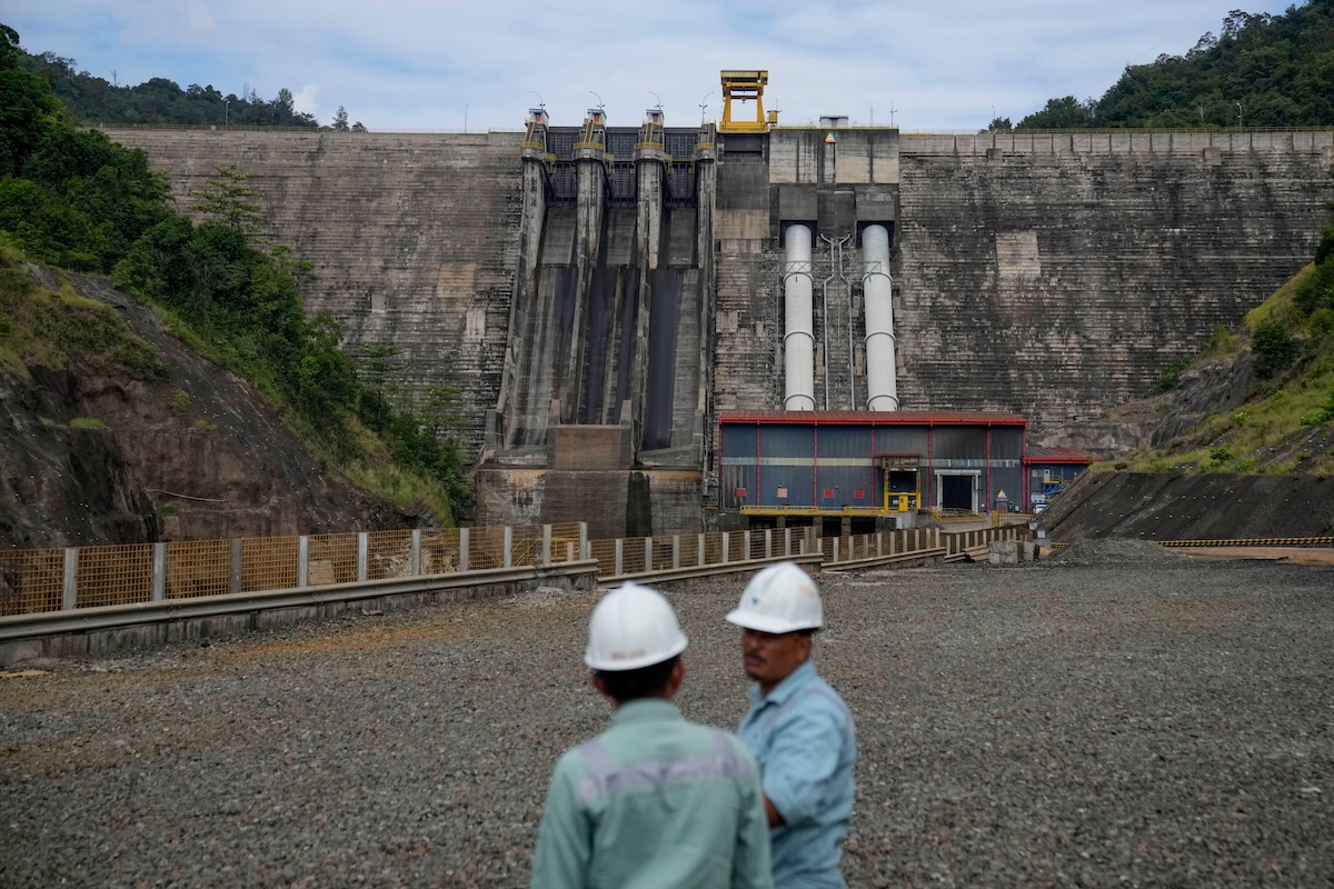 Two workers in hard hats stand in front of a large concrete dam