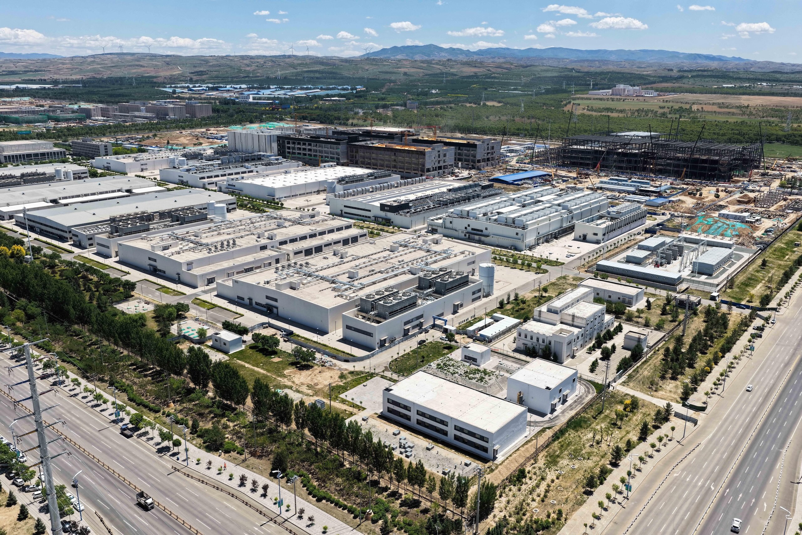 An aerial view of a large data centre with construction sites, and surrounding roads under a clear blue sky.