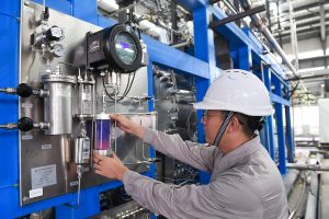 A technician adjusts a glass cylinder containing purple and pink liquid on a complex industrial control panel in a factory.