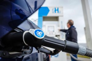 A close-up of a black hydrogen fuel nozzle with an H2 logo, with a blurred person operating a hydrogen refueling station in the background outdoors.