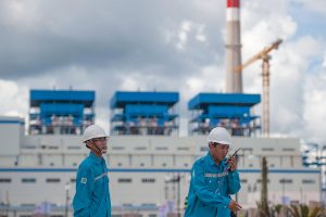 Two workers in blue uniforms and white hard hats stand before a large plant