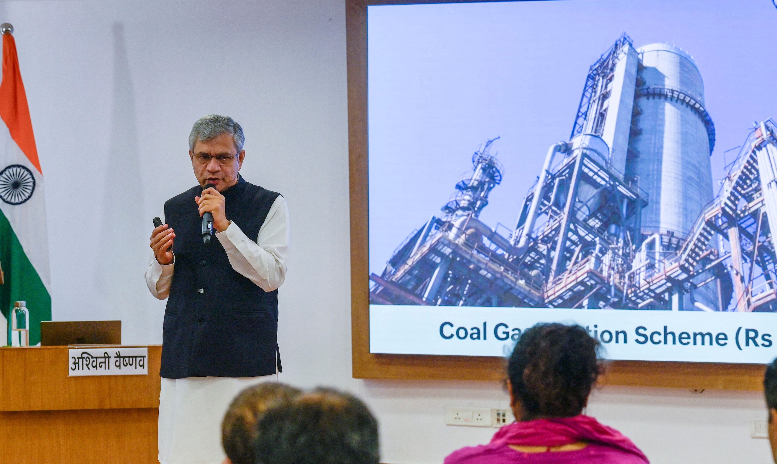 A man speaks at a podium before a large screen displaying a coal gasification plant