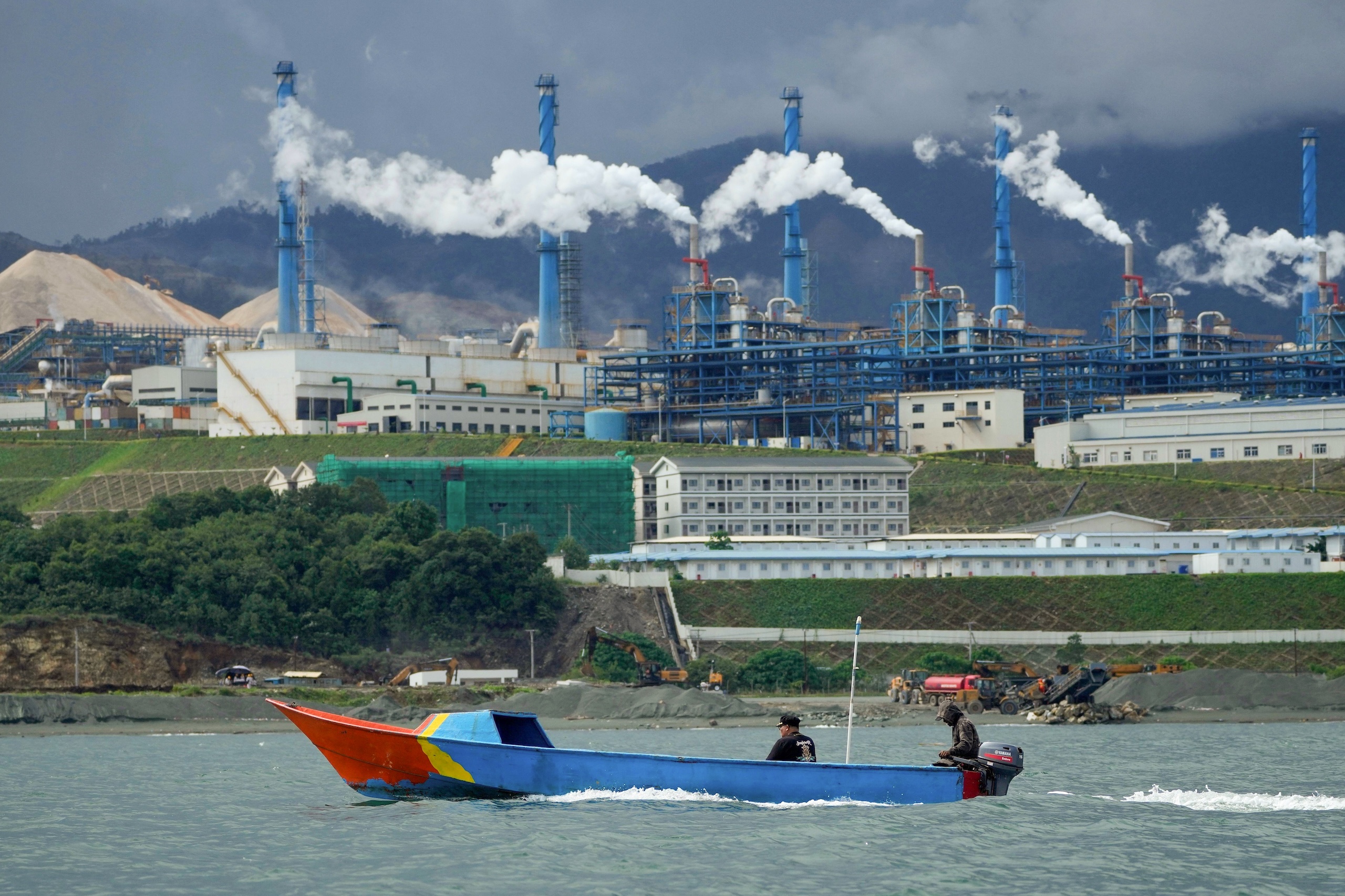 A boat with two people travels across the water in front of a large industrial factory complex with tall, smoking chimneys.