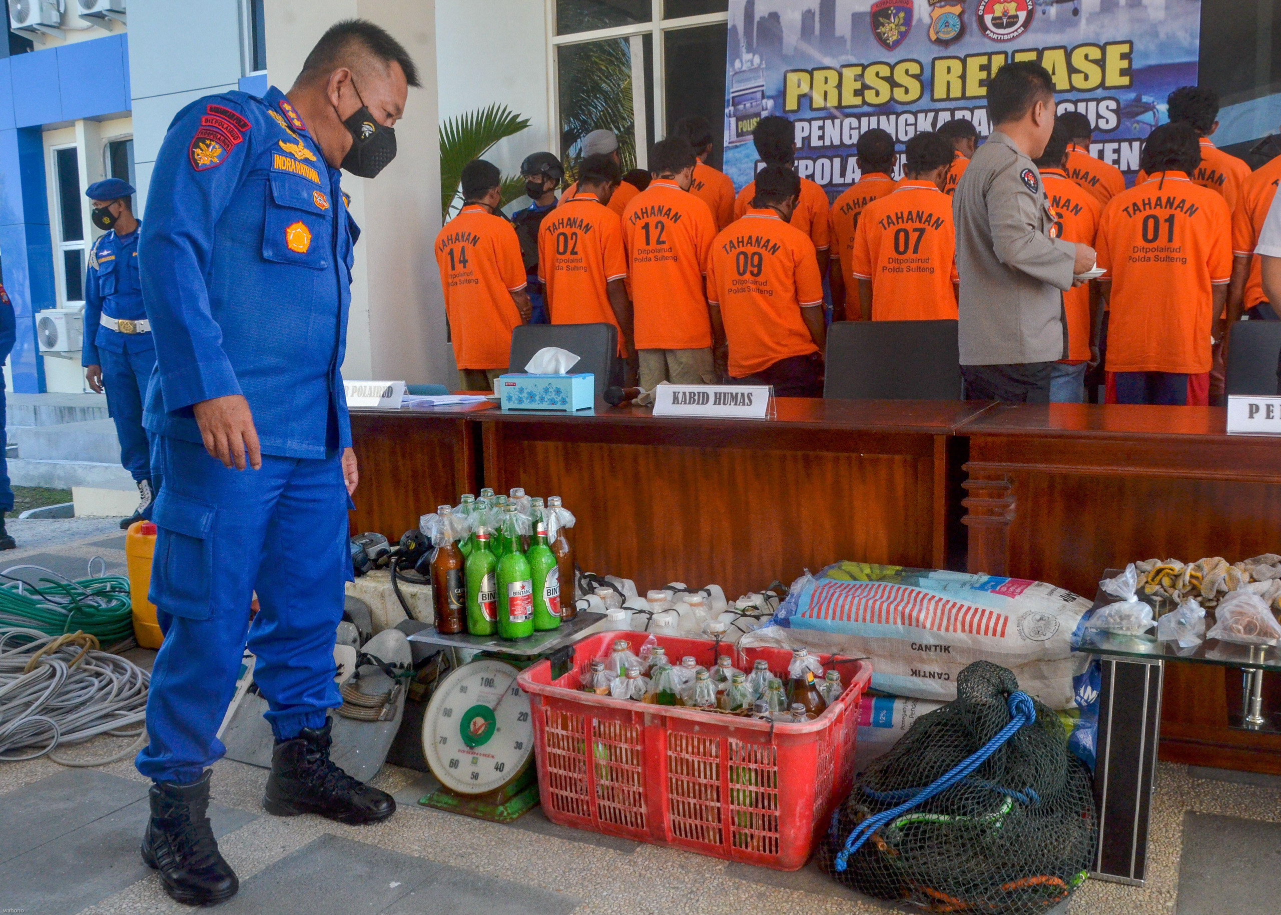 An official looks at items on the ground, including bottles, while men in orange stand in line facing away
