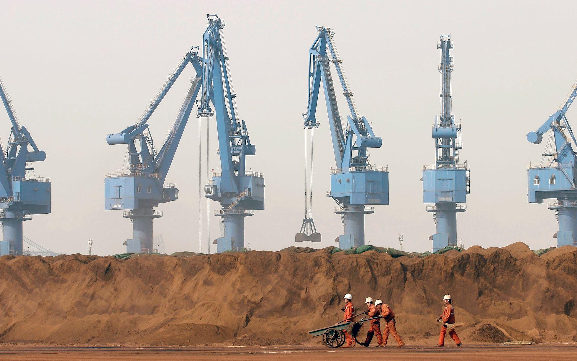 Four workers walk past a large pile of iron ore powder in front of several industrial port cranes.