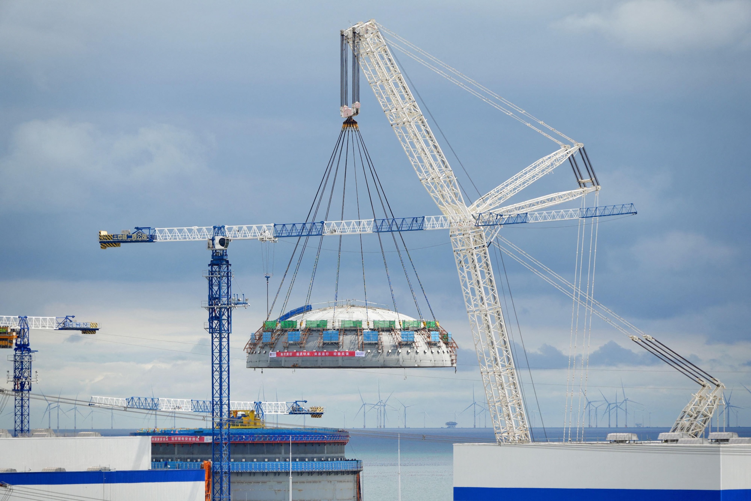 A large white crane lifts a massive, circular metal dome structure at a construction site near the ocean