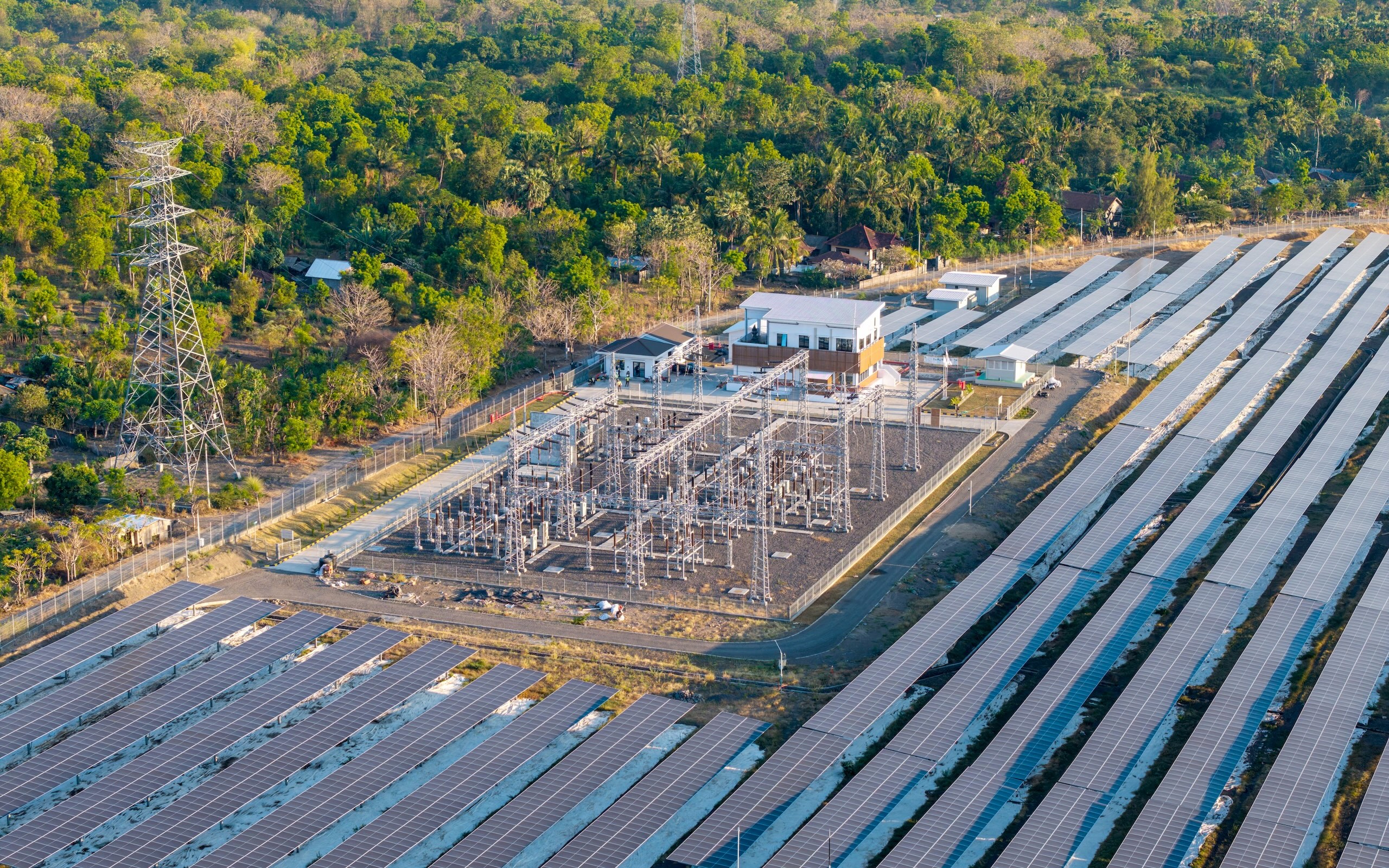 Aerial view of a solar power plant with solar panels in rows, surrounded by greenery and a power substation in the center.