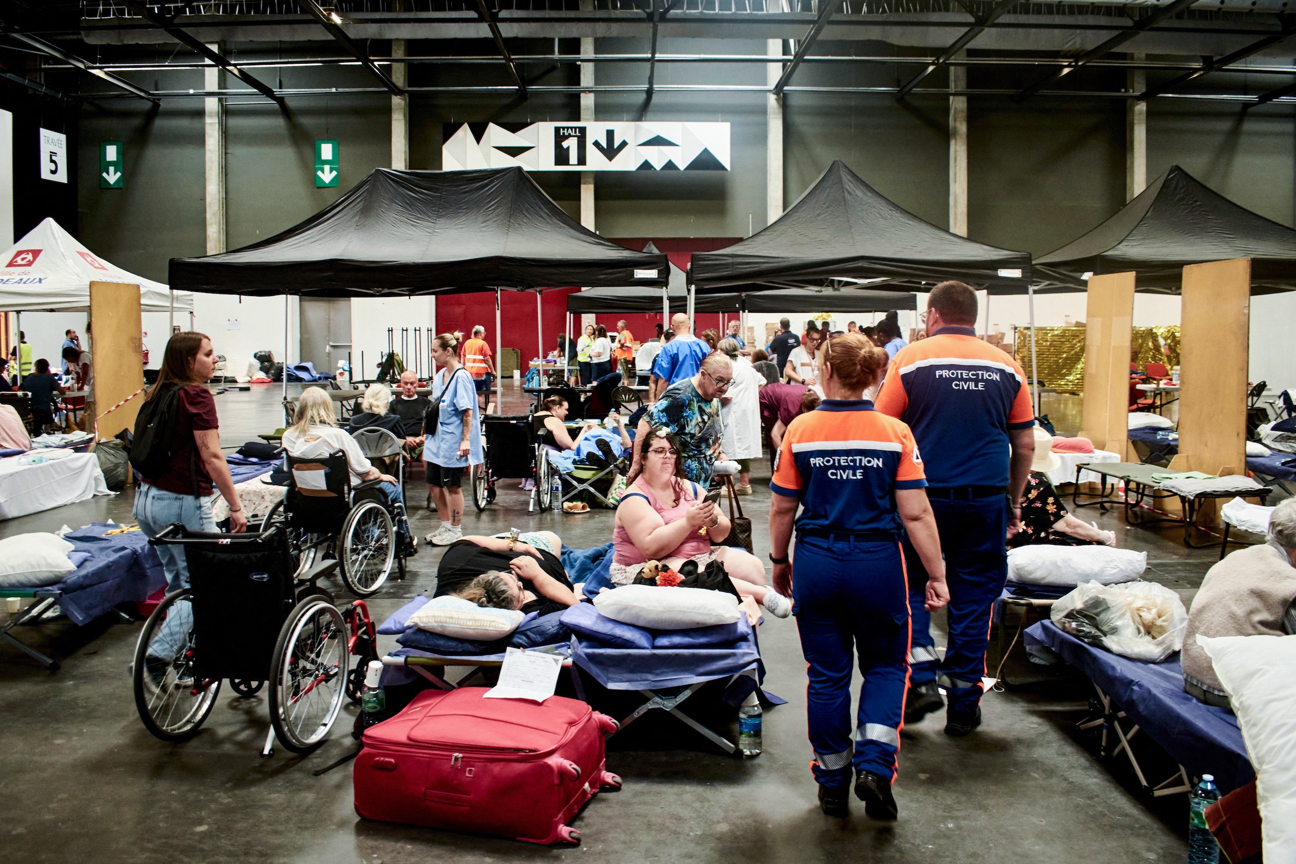 Two Protection Civile workers walk through a large hall filled with cots, wheelchairs, and people resting under black tents in a temporary shelter.
