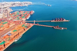 Aerial view of a coastal industrial port featuring large piles of raw materials, cargo ships docked at piers