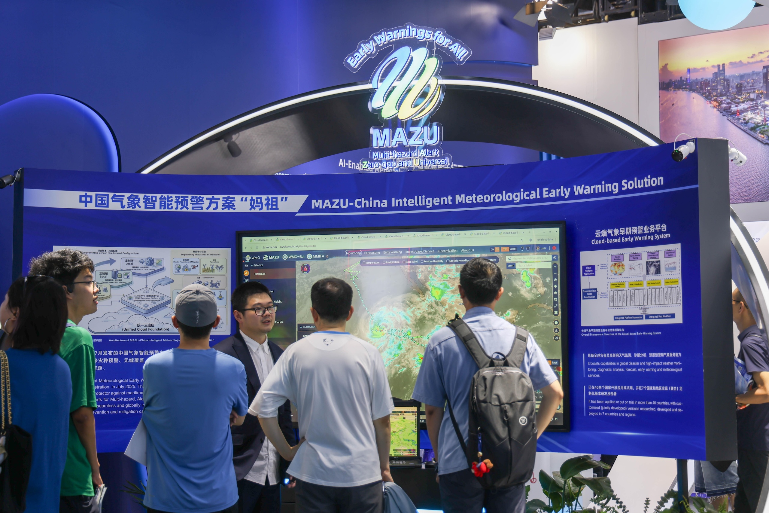 A group of people stands before a large digital display showcasing the MAZU-China Intelligent Meteorological Early Warning Solution at a show room.