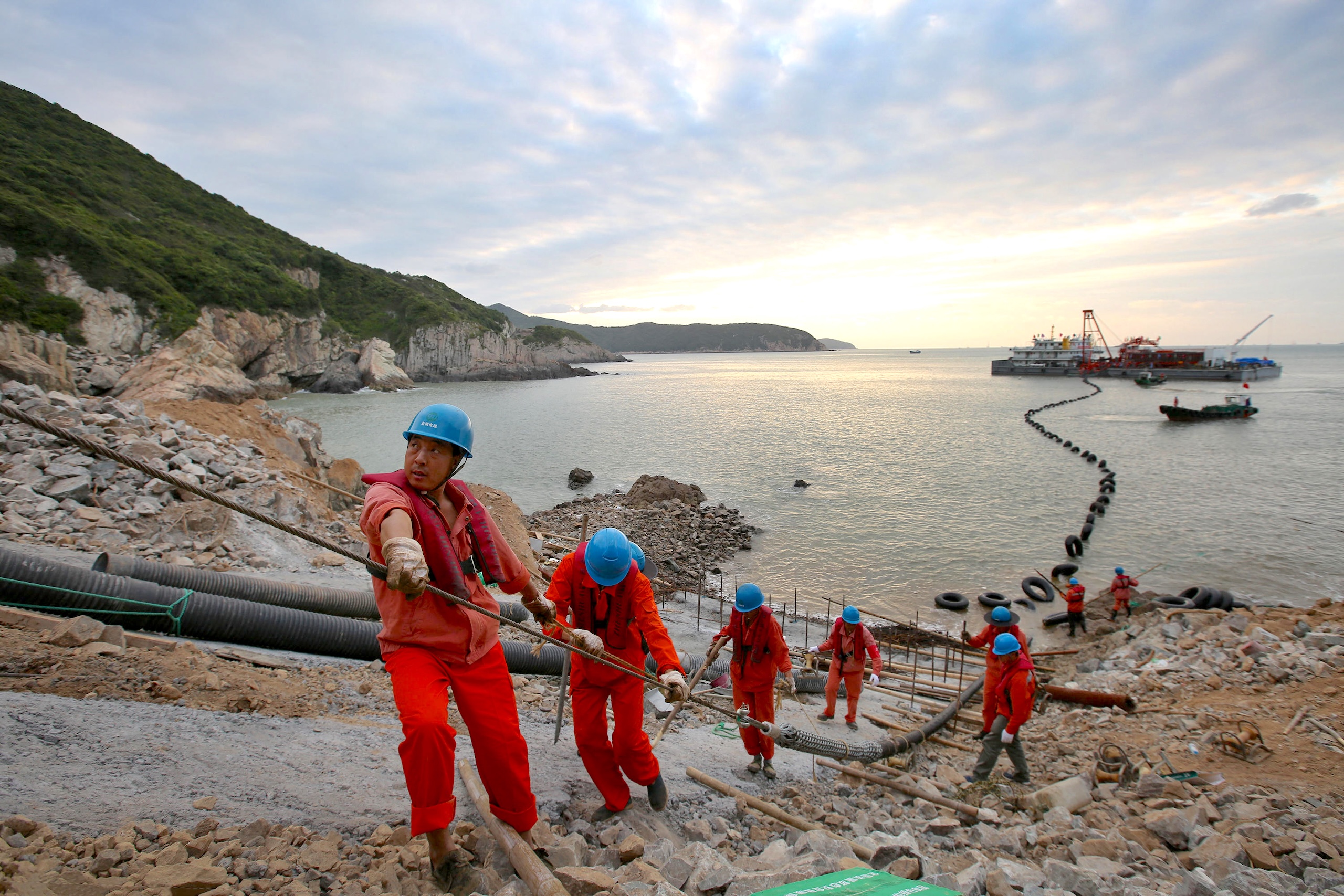 Workers in orange uniforms and blue hard hats pull a thick cable on a rocky shore, with a large industrial vessel visible in the water behind them.