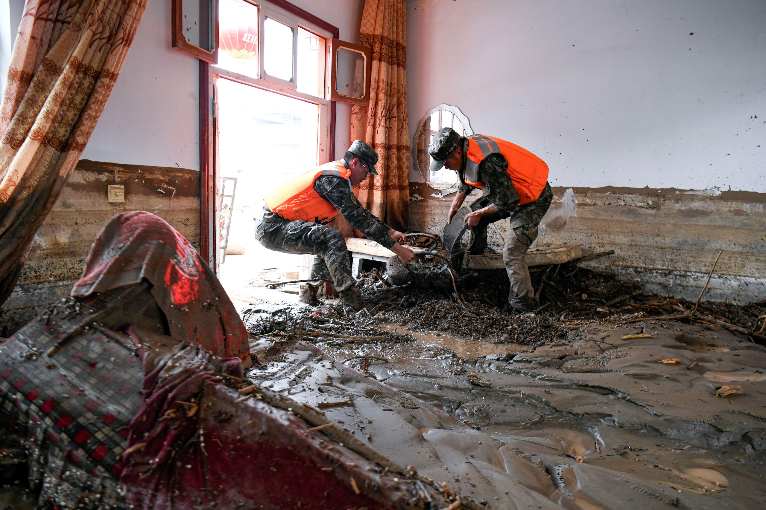 Two people in life vests clear mud and debris from the interior of a room flooded with thick, dark brown sediment.