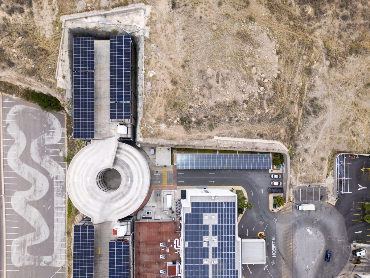 Aerial view of a hospital facility equipped with extensive solar panel arrays on its rooftops and parking structures, situated in an arid landscape in