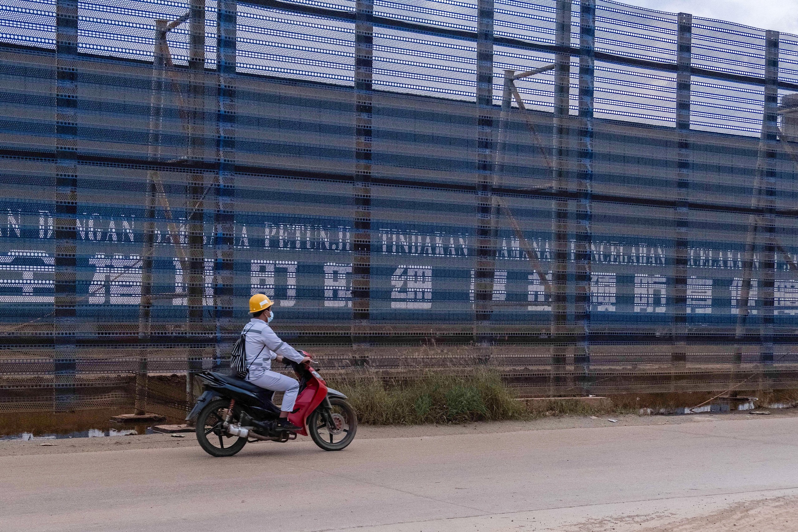 A person in a yellow hard hat rides a red motorbike past a big blue metal mesh fence